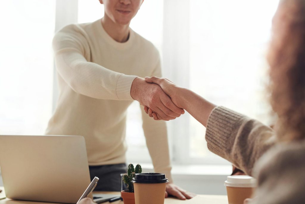 Two people shaking hands in a modern office setting, symbolizing collaboration and partnership, with a laptop and coffee cups on the table, relevant to online marketing and business strategies.
