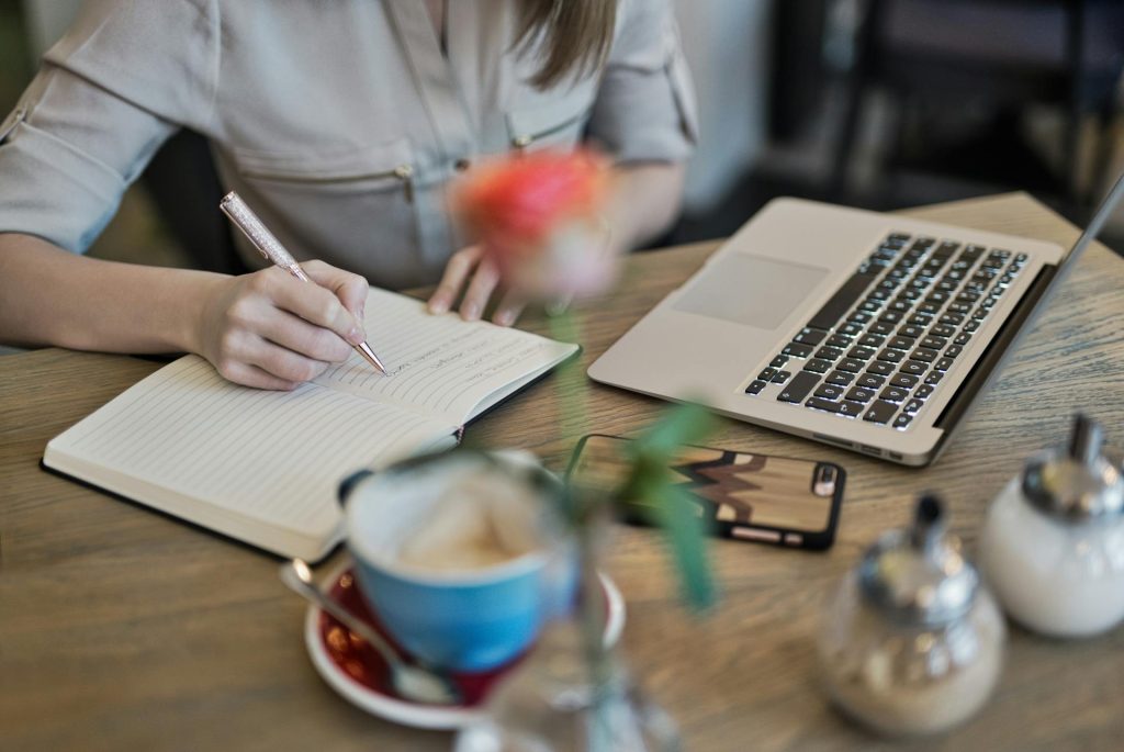 Person writing in a notebook with a laptop, coffee cup, and mobile phone on a table, illustrating online marketing and affiliate marketing strategies.